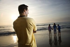 Caucasian mid-adult man standing and watching mid-adult woman with children on beach.