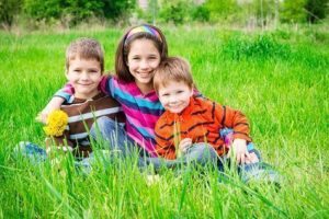 Group of smiling kids with dandelions sitting on the green grass meadow